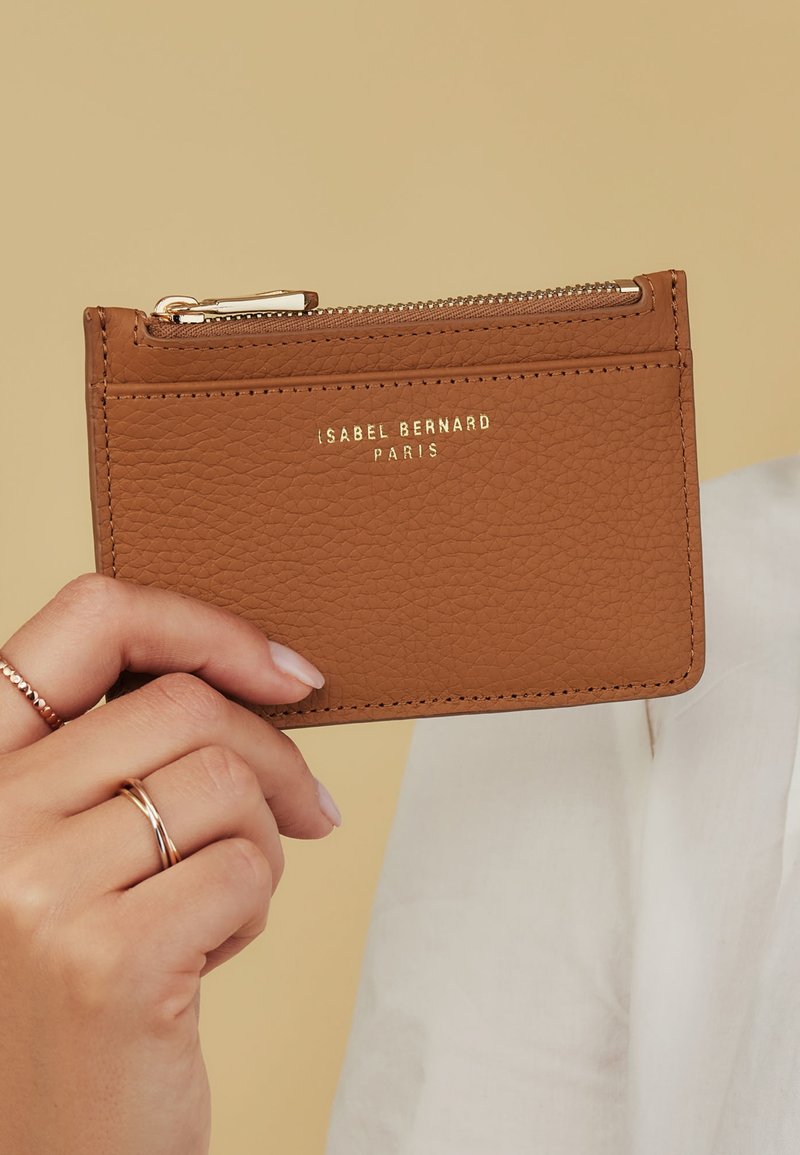 Tan leather cardholder with a textured surface, gold embossed logo, zippered compartment, and multiple card slots on the front.