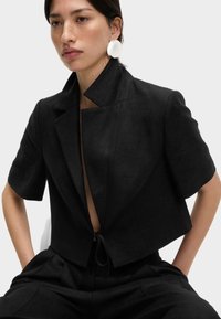 Schwarze, kürzere Blazerjacke mit kurzen Ärmeln, breitem Kragen und einem Kordelzug in der Taille; aus strukturiertem Stoff hergestellt.