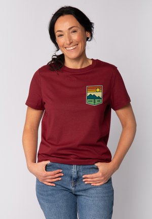 T-shirt en coton bourgogne avec un écusson graphique représentant des montagnes, un soleil et le texte "GOOD VIBE". Manches courtes et col rond.