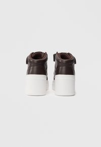 Bruine leren high-top sneakers met dikke witte platformzolen, bekeken vanaf de achterkant op een effen lichte achtergrond.