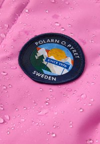 Vaatepunainen vedenpitävä kangas, jossa on pyöreä tummansininen paikka vuoristolla, auringolla ja teksteillä "POLARN O. PYRET," "SINCE 1976" ja "SWEDEN" pisaroiden kanssa.