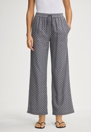 Graue gemusterte weit geschnittene Hose mit elastischem Bund und Taschen; der Stoff hat ein geometrisches Design. Kombiniert mit schwarzen Flip-Flops.