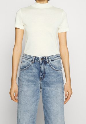 Person trägt ein schlichtes weißes kurzärmeliges Oberteil, das in eine hoch taillierte hellblaue Jeans gesteckt ist, vor einem schlichten Hintergrund.