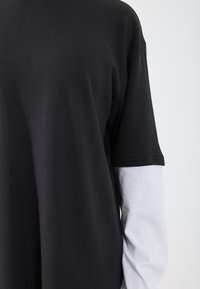 Czarna, prążkowana koszulka z krótkim rękawem, nałożona na białą koszulę z długim rękawem. Widoczna tekstura i kontrastujące kolory tworzą głębię.