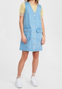 Ärmelloses Denimkleid in Hellblau, mit Knopfleiste vorne, zwei vorderen Taschen und bequemer Passform. Darunter wird ein gelbes, gemustertes Oberteil getragen.