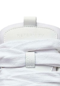 Valkoinen lenkkari, jossa on teksturoitu kangasosa, litteät valkoiset nauhat ja vahvistettu nahkakiila, jossa on kohokirjaimin merkintä "RETERNITY".