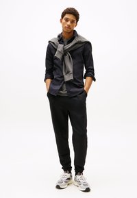 Jovem de pé com as mãos nos bolsos, vestindo uma camisa preta por cima de uma t-shirt cinza, sweatshirt cinza pendurada nos ombros, calças pretas e ténis brancos.