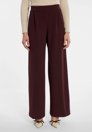 Vrouw draagt donker bordeauxrode broek met wijde pijpen en hoge taille met plooien aan de voorkant en beige puntige schoenen met zwarte riemen.