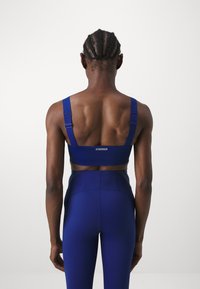Sinine sportlik rinnahoidja laiente rihmadega, tekstuurse kujunduse ja perforatsioonidega. Tagaküljel on logo "STRONGER".