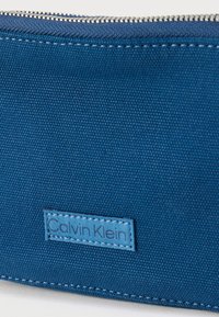 Μπλε υφασμάτινη θήκη με λευκό φερμουάρ και ραμμένη ετικέτα με το λογότυπο Calvin Klein κοντά στην κάτω άκρη, που δείχνει λεπτομερή υφή και ραφές.