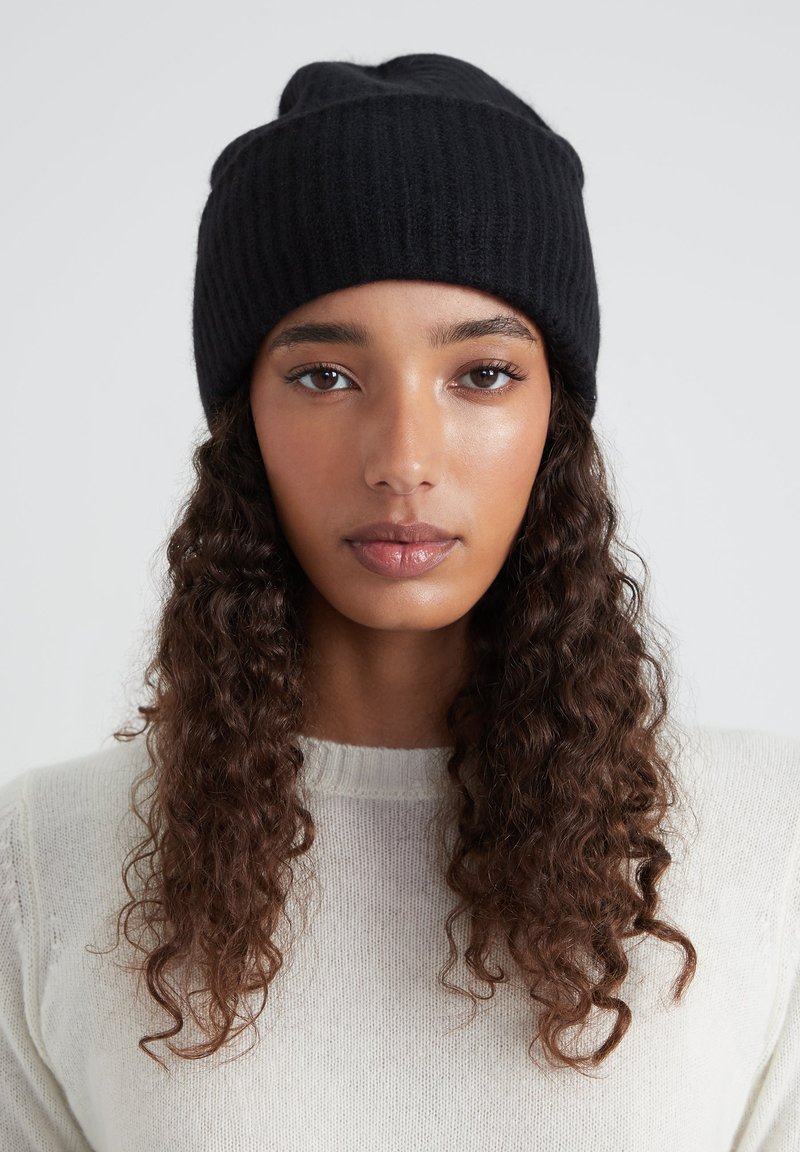 Cappello a cuffia nero in maglia a coste con risvolto, realizzato in materiale morbido, indossato su una modella con capelli ricci, su uno sfondo neutro.