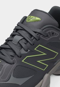 Κοντινό πλάνο ενός αθλητικού παπουτσιού σε μαύρο και γκρι χρώμα με πράσινες λεπτομέρειες, διχτυωτές επιφάνειες, χοντρές κορδόνια και την ένδειξη "9060" στη γλώσσα.