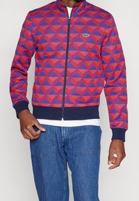 Veste à motif géométrique rouge et violet avec fermeture éclair, col montant et poignets côtelés bleu marine, portée sur un jean en denim.