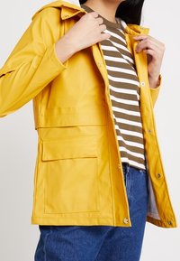Veste imperméable jaune avec capuche, boutons et poche frontale, portée sur une chemise rayée marron et blanche, associée à un jean bleu.