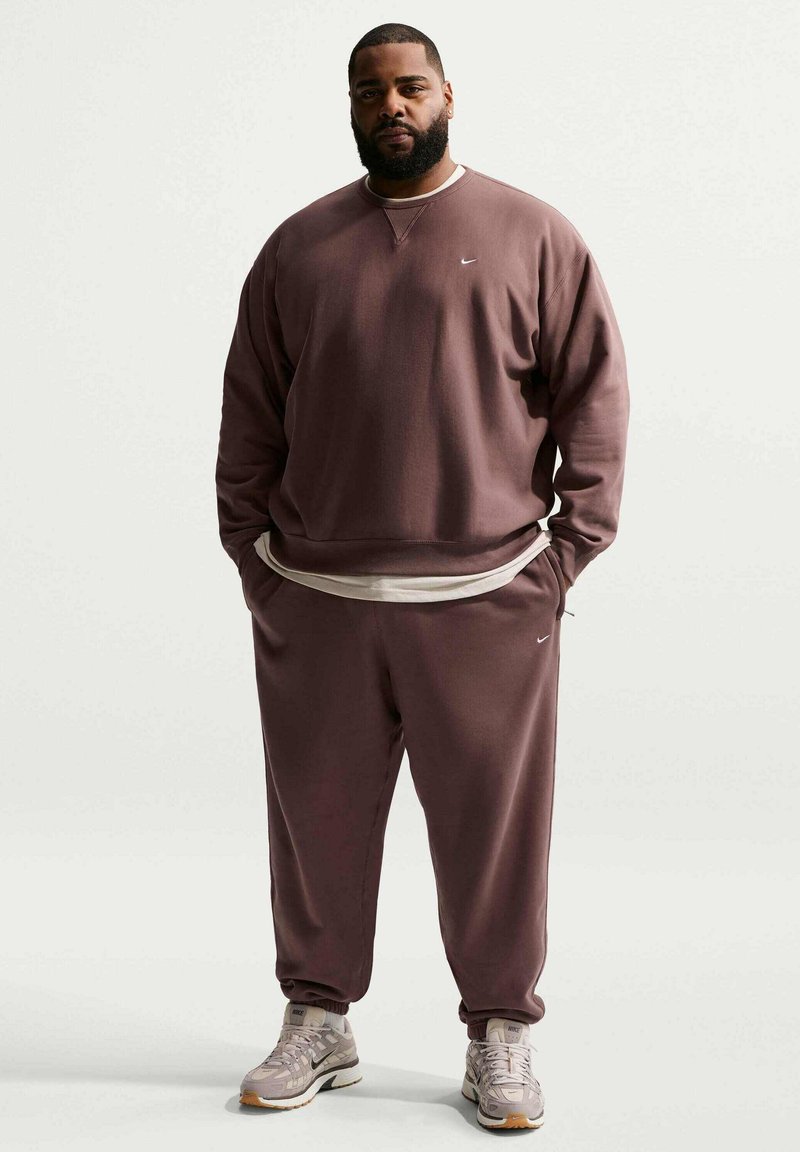 Man staat, draagt een losse bruine Nike-trui en bijpassende joggingbroek, beige ondershirt en grijze sneakers tegen een effen achtergrond.