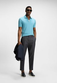 Light blue polo shirt with a zip neck, grey patterned pants, and dark shoes. Model holds a navy blazer, wearing sunglasses.