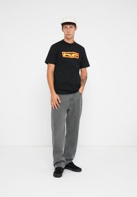Black cotton t-shirt with bold yellow and red logo, paired with gray denim pants. Model wears black sneakers and a white cap. Neutral background.