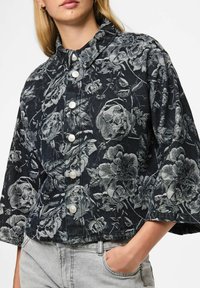Vrouw in een cropped zwarte jas met lange mouwen, voorzien van een grijs bloemenpatroon en witte knopen, gecombineerd met lichtgrijze jeans, hand in de zak.