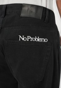 Černé džíny s koženou nášivkou na pásku a bílým vyšitým textem "No Problemo" na zadní kapse.