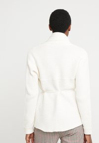 Cárdigan blanco de punto con textura de canalé, con el frente abierto y un lazo a la cintura, y mangas largas, combinado con pantalones de cuadros.