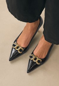 Crne balerinke od lakirane kože šiljastog vrha s velikom zlatnom lancem kao ukrasom, nošene s crnim skraćenim hlačama na bež podu.