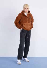 Brun fleece-hoodie med kvartsdress, framficka och svarta jeans. Modellen bär vita sneakers med lila ränder. Enkel bakgrund.