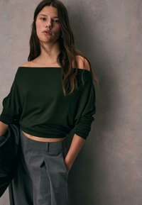 Top s odhalenými ramenami, tmavozelený, vyrobený z mäkkého materiálu, v kombinácii s vysokoklasickými, sivými, prispôsobenými nohavicami; model stojí na neutrálnom pozadí.