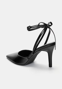 Scarpa nera in ecopelle con tacco alto, punta appuntita, cinturino sottile alla caviglia e un fiocco decorativo sul retro. Texture liscia, tacco affusolato.