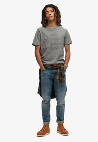 Grå bomulds t-shirt med præget logo, blå denim jeans, flannel skjorte bundet om livet og brune snørebåndsstøvler med hvide såler.