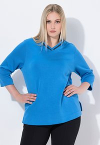 Top azul brillante de manga larga con escote en V y detalles en negro. Textura suave y corte holgado, con aberturas laterales para mayor comodidad.