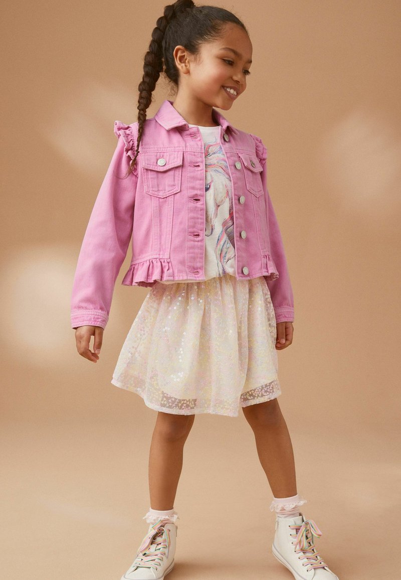 Veste en denim rose avec des épaules volantées sur une chemise blanche à motif coloré. La jupe est transparente avec des sequins et une finition en dentelle. Baskets blanches.