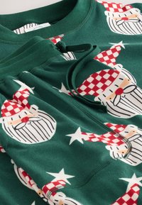 Zelená flaušová súprava na voľný čas s opakujúcim sa vzorom tváre Santa Clausa a hviezd, s červenými károvanými klobúkmi a vyšívanými detailmi.