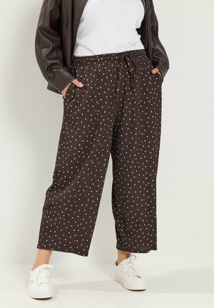 Persona que lleva pantalones sueltos marrones con lunares, zapatillas blancas, camiseta blanca y chaqueta de cuero marrón oscuro con las manos en los bolsillos.