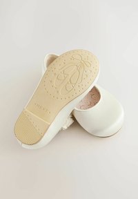 Des ballerines blanches présentent une finition satinée lisse, des accents floraux et une semelle en caoutchouc texturée avec des détails de design de ballet.