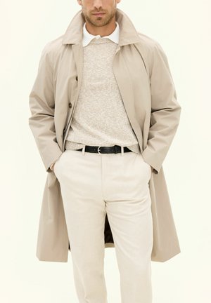 Trench coat beige con colletto a pistagna, maglione beige a fantasia e pantaloni chiari, fissati con una cintura nera.