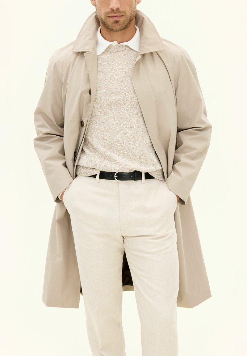 Trench coat beige con colletto a pistagna, maglione beige a fantasia e pantaloni chiari, fissati con una cintura nera.