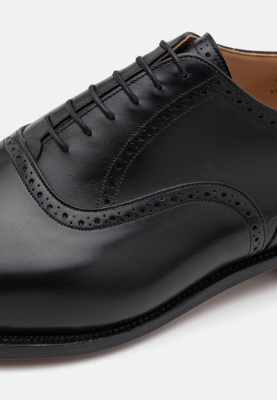 Scarpa da uomo in pelle nera con finitura liscia, presenta accenti perforati e una punta arrotondata, allacciata con cinque passanti. Cuciture dettagliate presenti.