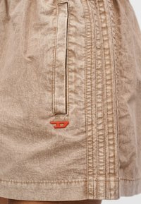 Ανοιχτό καφέ ύφασμα με κάθετη ραφή και στενή σχισμή τσέπης, με μικρό κόκκινο λογότυπο Diesel κοντά στο κάτω άκρο.