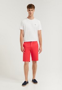 Röda bomullsshorts med klassisk passform, i kombination med en ljus vit t-shirt och marinblå espadrillos, stående mot en neutral bakgrund.