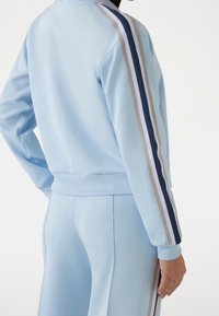 Ljusblå träningsdräkt med ribbade muddar. Har tre vertikala ränder i mörkblått, grått och vitt längs ärmarna. Mjuk textur.