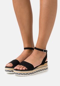 Černé semišové espadrilové sandály s širokým předním páskem, páskem kolem kotníku se sponou a vícvrstvou tkanou platformou, která kombinuje černé a béžové akcenty.