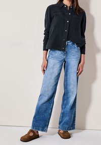 Chemise noire texturée à boutons avec poches, associée à un jean bleu clair à jambes larges et des sabots en daim marron. Design simple, coupe décontractée.