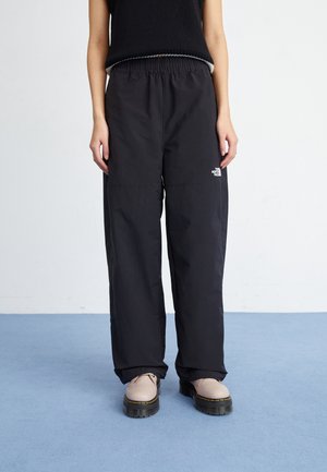 Osoba ubrana w czarne luźne spodnie z logo The North Face i beżowe sznurowane buty na niebieskiej podłodze na tle białej ściany.