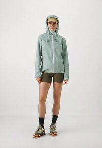 Vaaleanvihreä vedenpitävä takki, jossa on kokopitkä vetoketju, säädettävä huppu ja kiristysnyörit. Yhdistetty oliivinvihreiden shortseihin ja lenkkitossuihin.