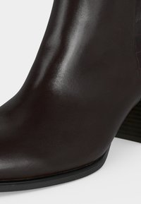 Gros plan d'une botte cheville en cuir noir avec une finition lisse et un talon bas empilé sur un fond blanc.