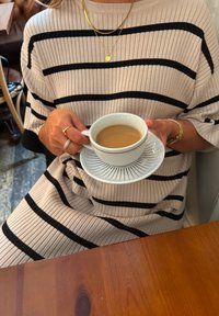 Vit keramikmugg med fat, innehållande en brun dryck. Randig beige och svart ribbad tröja med trekvartsärmar, guldsmmycken synliga.