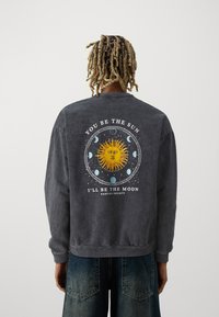 Person med blonda dreadlocks som bär en mörkgrå sweatshirt med sol- och månfaser grafik samt texten "Du är solen, jag är månen."