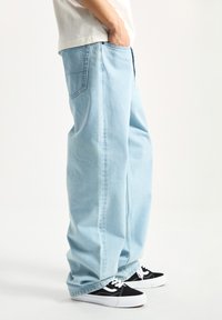 Jeans de mezclilla azul claro con un corte relajado, que presentan bolsillos traseros y una costura lateral, combinados con zapatillas negras y suelas blancas.