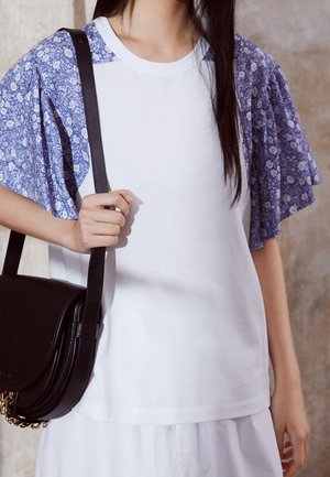 Persona con una camisa blanca con mangas cortas de flores azules y sostiene un bolso de hombro negro con detalle de cadena dorada.