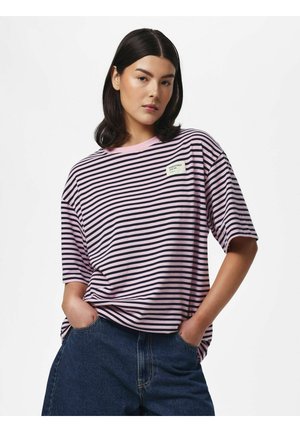 Jeune femme aux cheveux foncés portant un t-shirt rayé horizontal rose et noir et un jean bleu, les mains dans les poches.